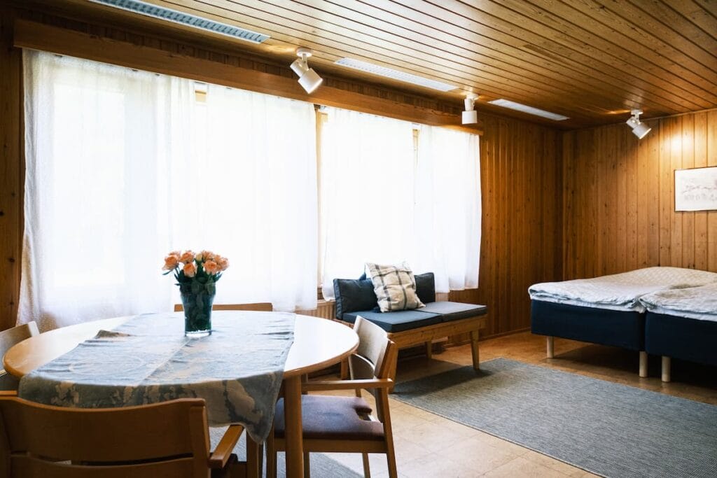 Ett rum med träpaneler och stora fönster, ett runt matbord med stolar och en vas med blommor, en liten soffa och en säng med kuddar och sängkläder.