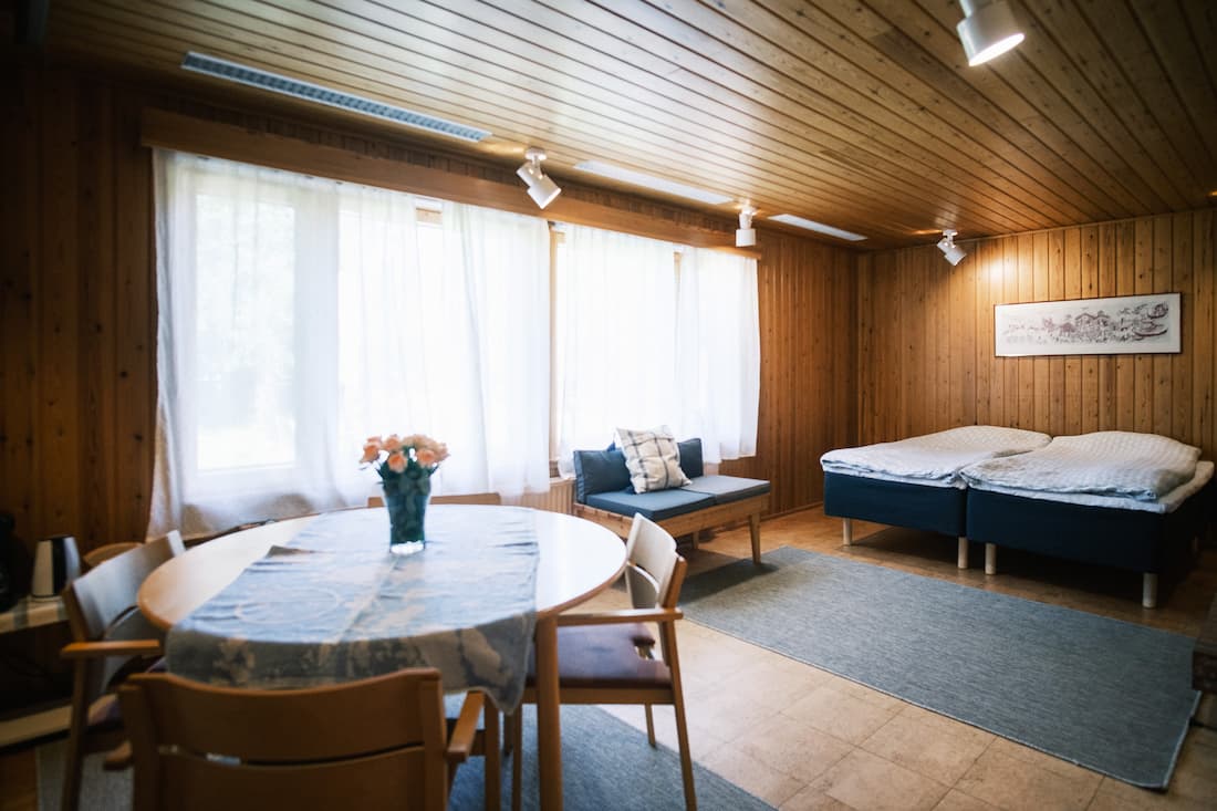 Rummet, som är klätt med träpaneler, har ett runt matbord med stolar, en liten soffa, två enkelsängar, stora fönster med tunna gardiner och en blå matta.