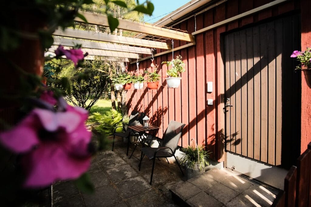 En uteplats med två svarta stolar och ett litet bord bredvid en röd träbyggnad; hängande blomkrukor och grönska syns i solljuset.