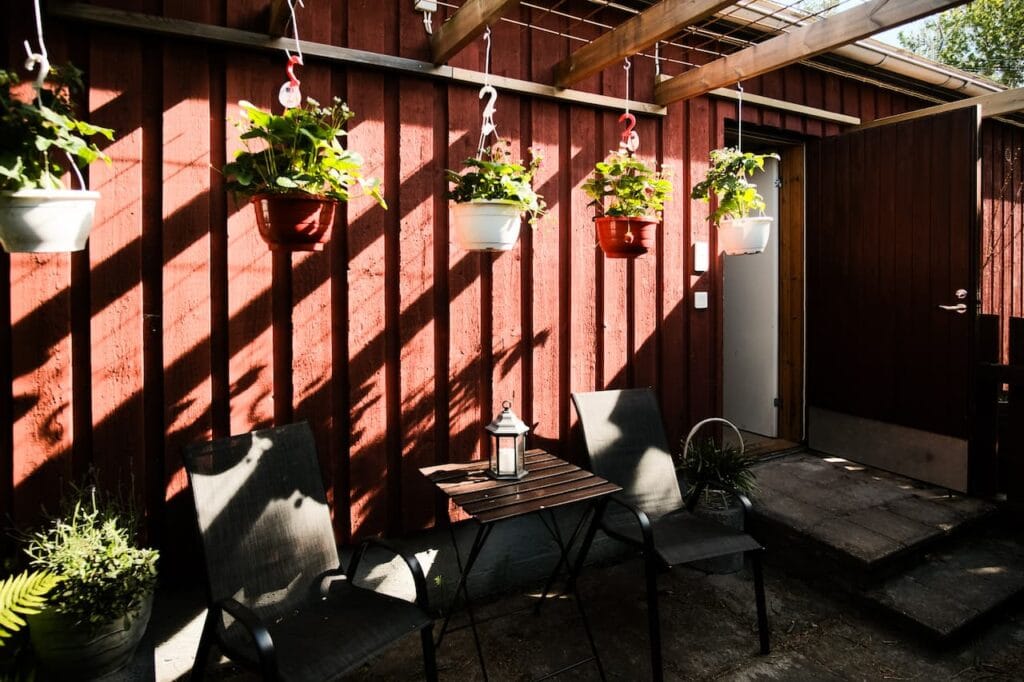 Två svarta stolar och ett litet bord med en lykta står på en uteplats intill en röd trävägg, med hängande krukväxter ovanför och solljus som kastar skuggor.