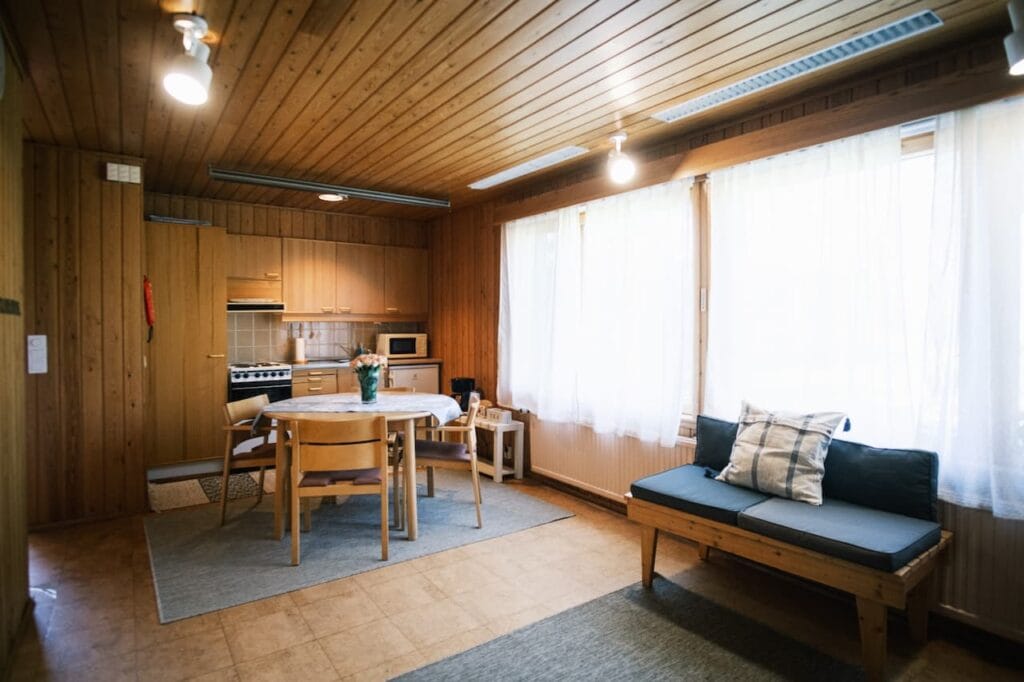 Ett kök och en matplats med träpaneler, ett runt bord, fyra stolar, en liten soffa, stora fönster med tunna gardiner samt grundläggande köksutrustning.