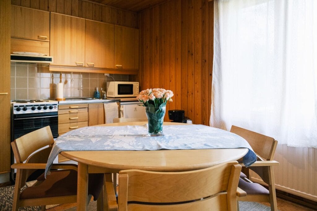 Ett litet kök med träskåp, ett runt bord med en blomsterdekoration i mitten, fyra stolar och naturligt ljus som strömmar in genom ett fönster med gardiner.