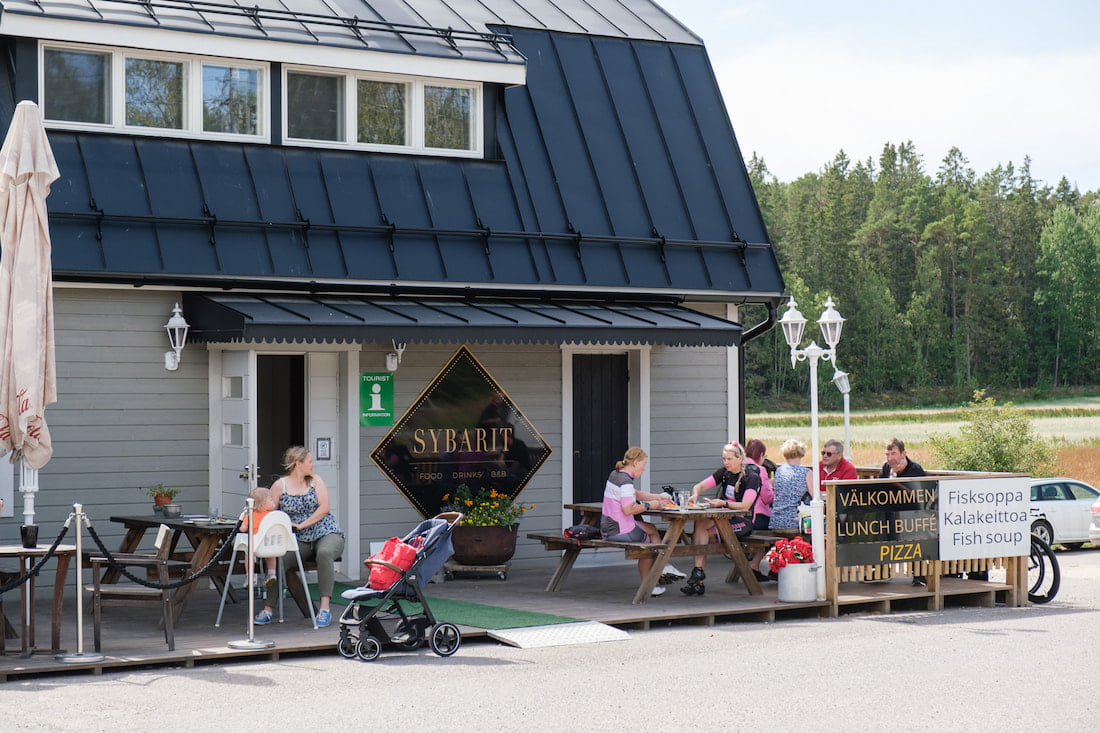Människor som äter vid utomhusbord framför en restaurang med en skylt där det står "sybarit.