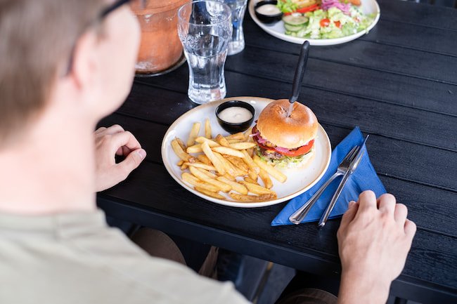 Person som sitter vid ett bord med en hamburgare, pommes frites och sallad redo att äta.