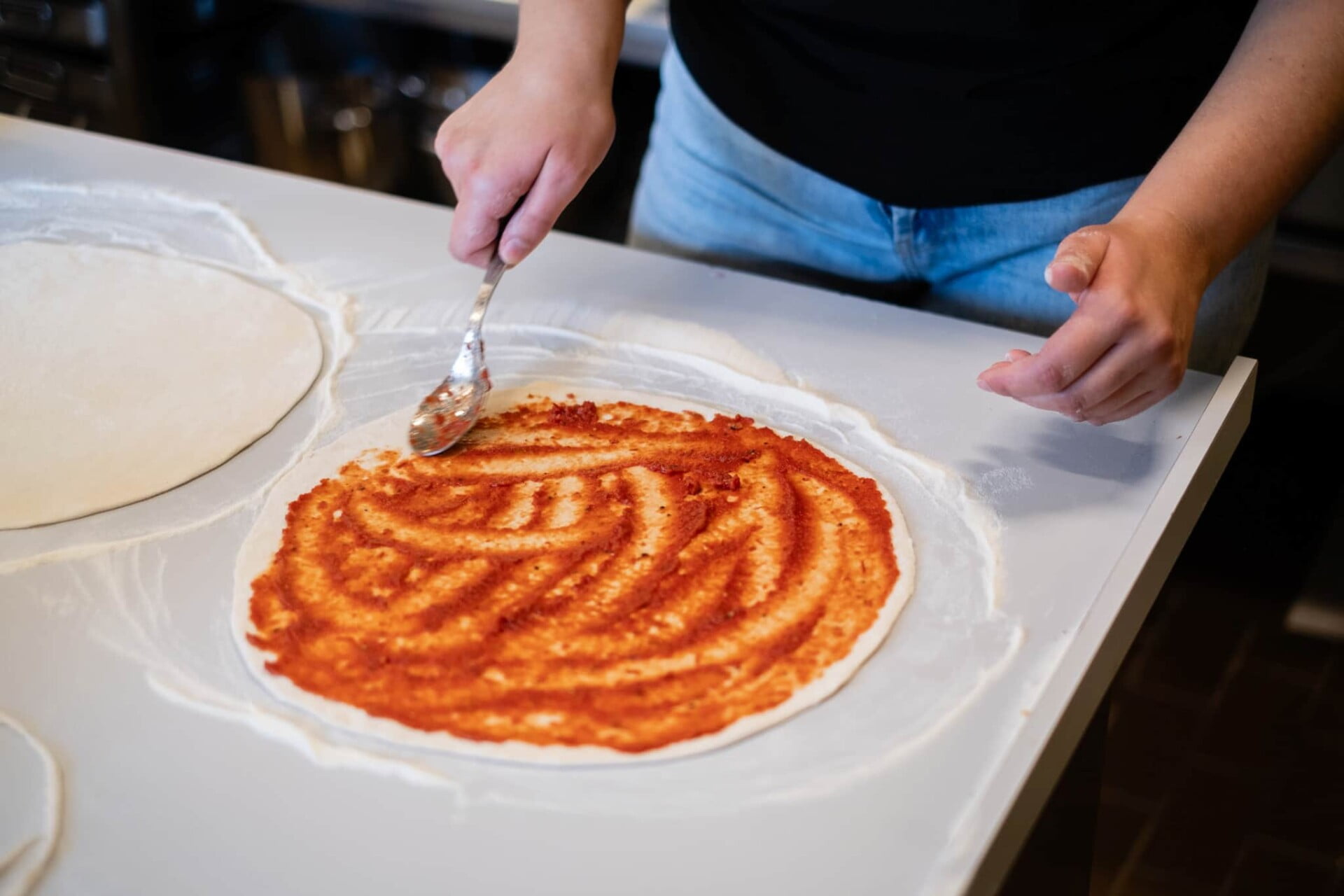 Person spreading tomato sauce on pizza dough with a spoon.