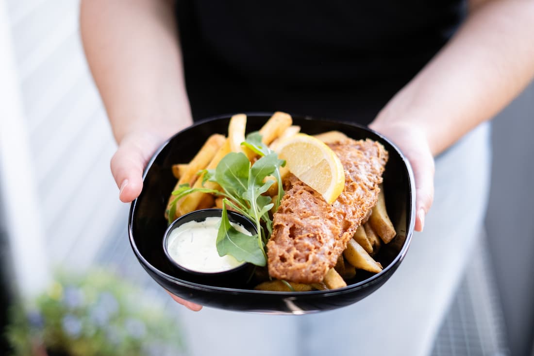 A person holding a plate of fish and chips with a side of tartar sauce and a lemon wedge.