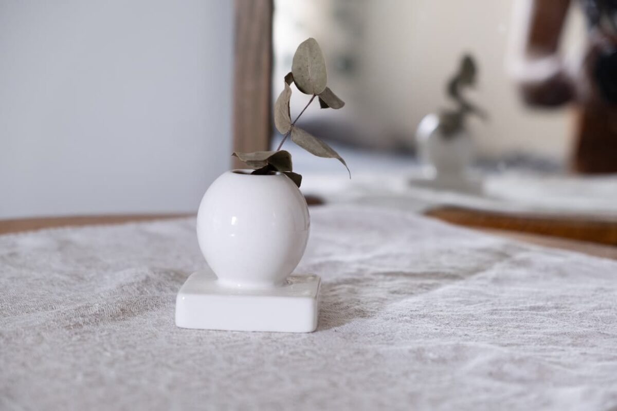 En enda eukalyptuskvist i en liten vit sfärisk vas på en texturerad tygyta med en suddig spegel i bakgrunden.