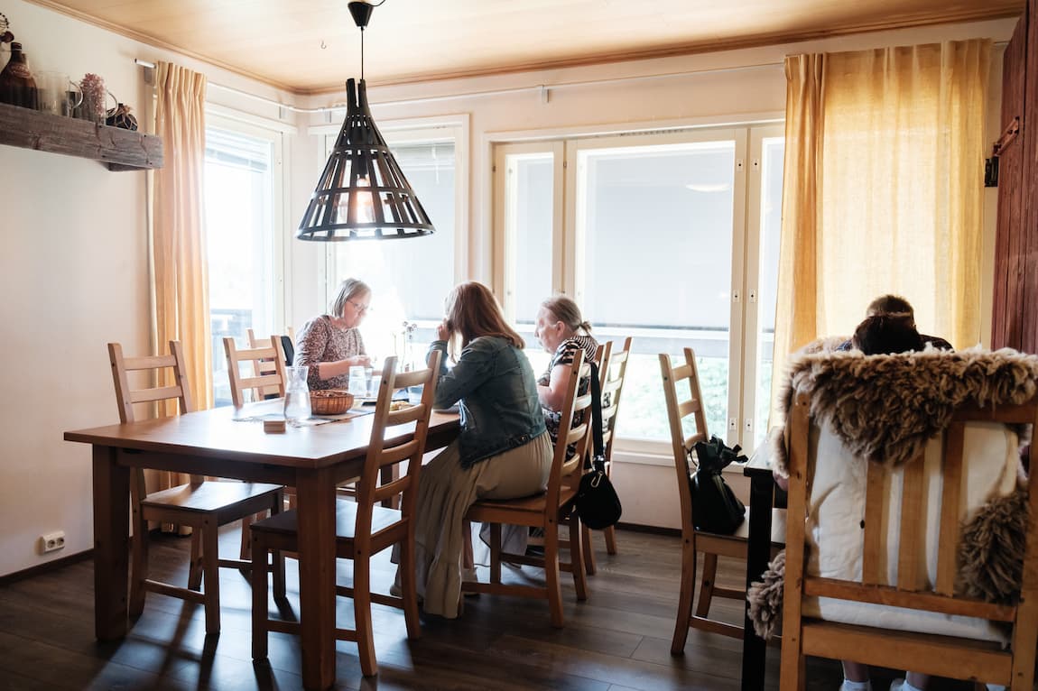 Tre personer njuter av en måltid tillsammans vid ett matbord i ett väl upplyst rum med balkong.