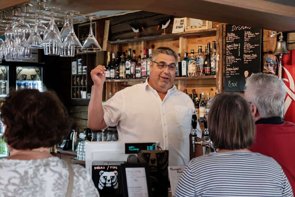 En man i vit skjorta talar till kunder vid en bardisk med drinkar i bakgrunden.
