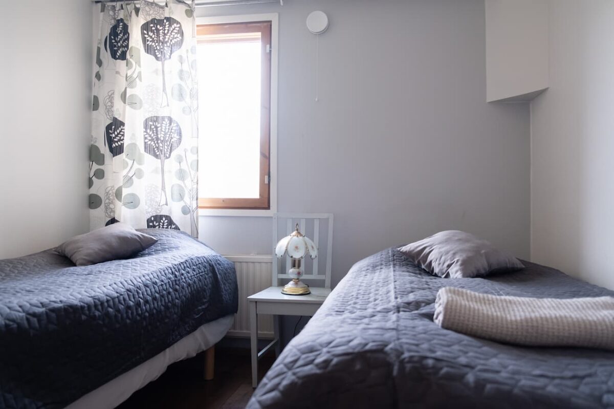 Två enkelsängar med blå täcken i ett litet, prydligt sovrum med fönster och nattduksbord med lampa och böcker.